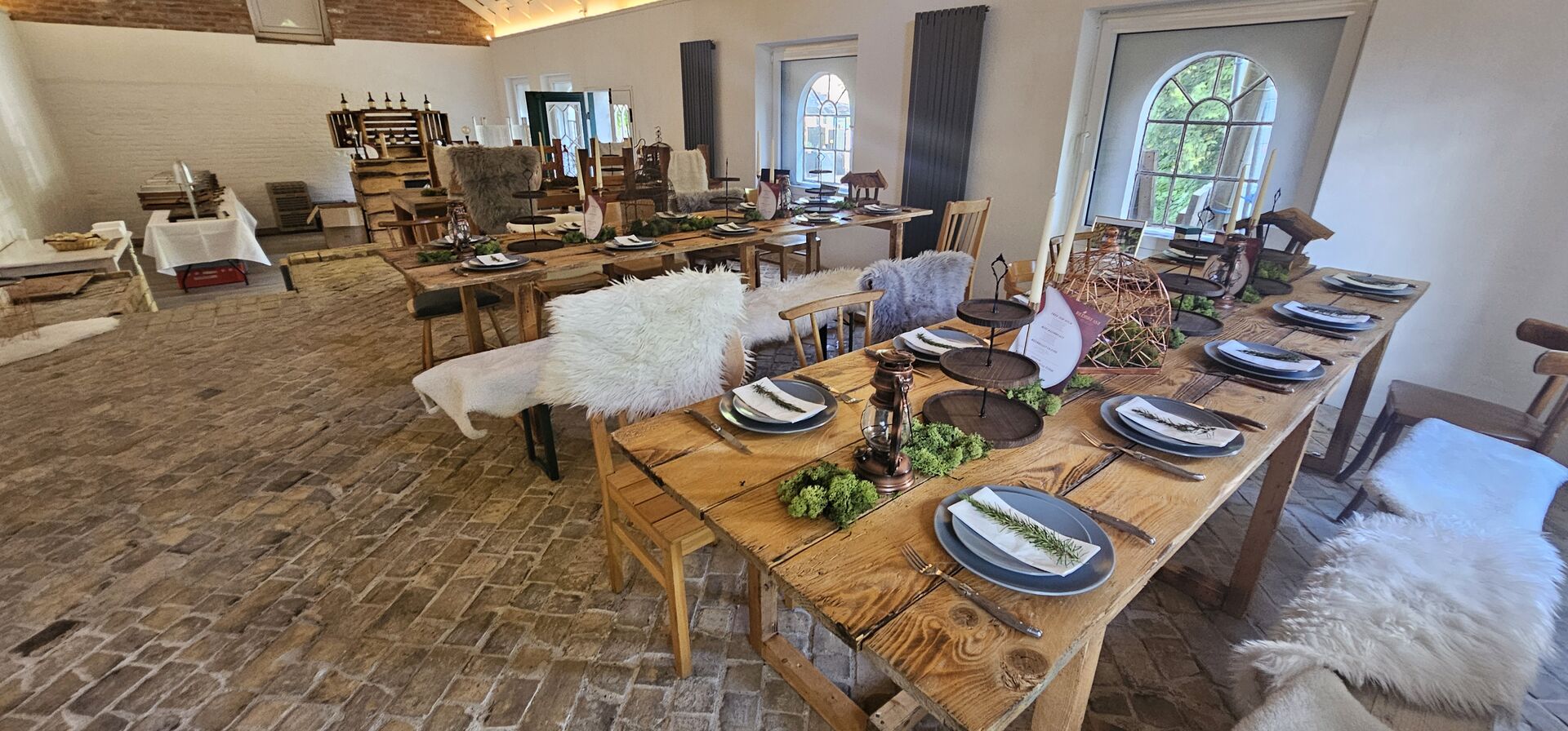 Scheune mit rustikalem Boden, Tischen, Stühlen und Bänken, auf Bänken und einigen Stühlen liegen Schaffelle
