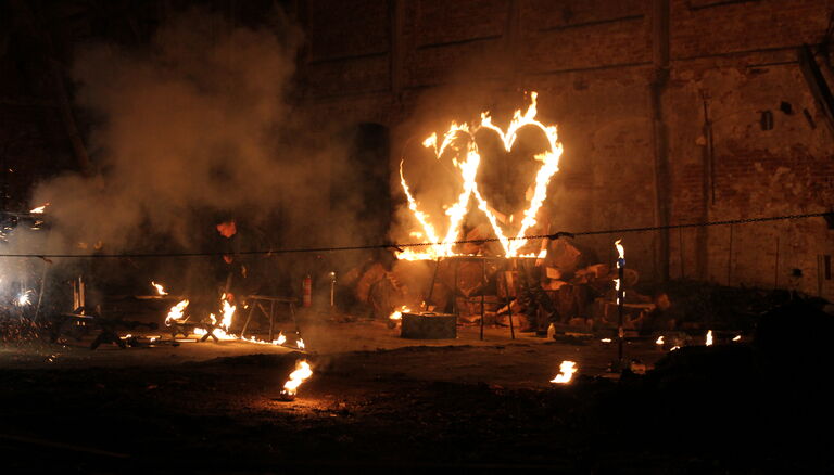 Rechts zwei brennende, herzförmige Metallrahmen. Links ein Mann, der mit Feuer interagiert. Mehrere Fackeln und brennende Schalen stehen um den Platz herum.