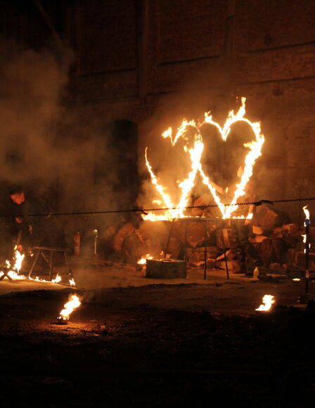 Rechts zwei brennende, herzförmige Metallrahmen. Links ein Mann, der mit Feuer interagiert. Mehrere Fackeln und brennende Schalen stehen um den Platz herum.