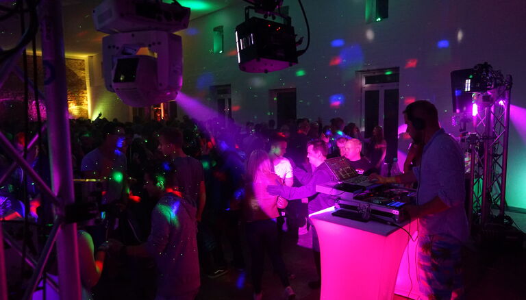 Rechts DJ Pult. Links und Rechts davon Gerüst mit Leuchten. Der Rest der Scheune ist voll mit Leuten.