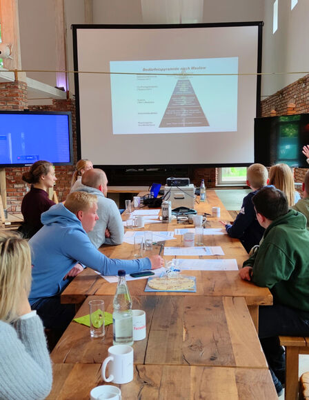 Seminar in Eventscheune, Teilnehmer sitzen an langem Tisch vor mobilen Bildschirmen. Aufgenommen vom Tischende