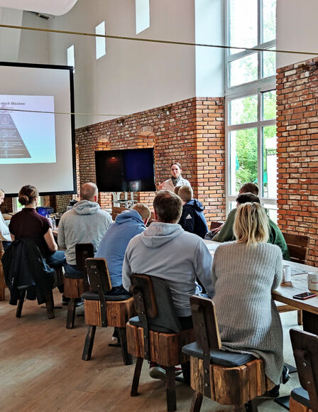 Seminar in Eventscheune, Teilnehmer sitzen an langem Tisch vor mobilen Bildschirmen. Aufgenommen von links-hinten