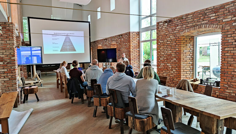 Seminar in Eventscheune, Teilnehmer sitzen an langem Tisch vor mobilen Bildschirmen. Aufgenommen von links-hinten
