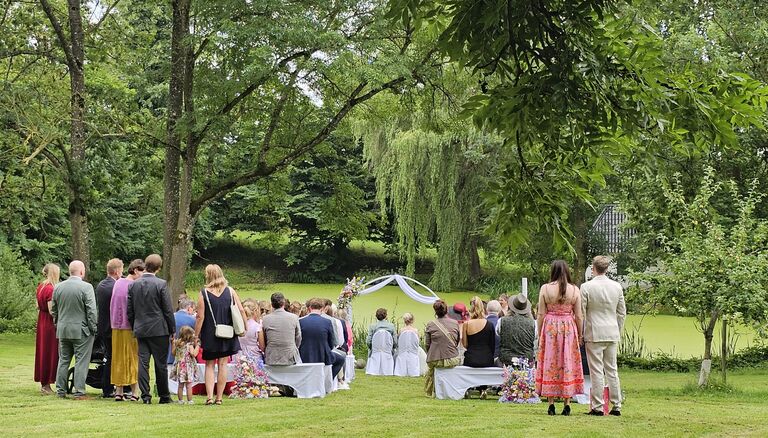 Hochzeitsgesellschaft am See. Das Paar schaut zum See. Die Gäste stehen auch mit dem Rücken zur Kamera. Im Hintergrund Bäume und ein Häuschen.