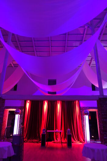 Scheune im Dunkeln, Kopfseite aus relativer näher fotografiert. Stoffbahnen hängen von Balken unter der Decke. Raum ist rot, pink und violett beleuchtet.