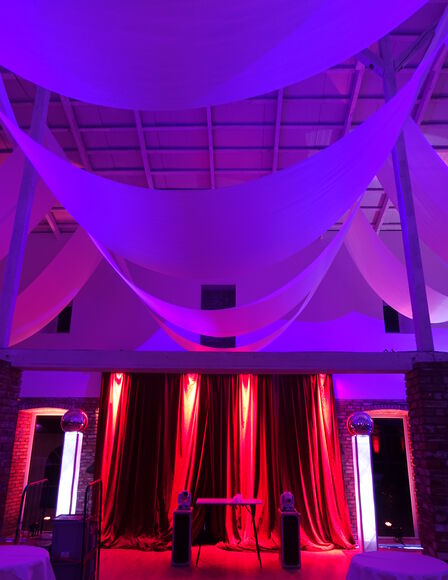 Scheune im Dunkeln, Kopfseite aus relativer näher fotografiert. Stoffbahnen hängen von Balken unter der Decke. Raum ist rot, pink und violett beleuchtet.