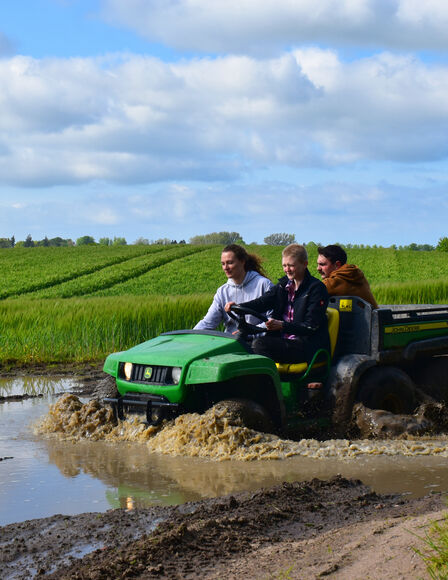 Eine Frau und zwei Männer fahren auf einem John Deere Gator durch eine matschige Pfütze