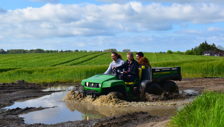 Eine Frau und zwei Männer fahren auf einem John Deere Gator durch eine matschige Pfütze