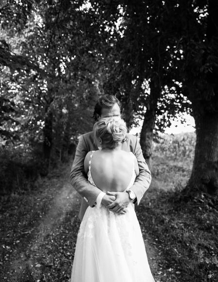 Bild schwarz-weiss, Hochzeitspaar umarmt sich vor Feldweg, Braut mit Rücken zur Kamera, Bräutigam mit Gesicht zur Kamera, sein Gesicht wird vom Kopf der Braut größtenteils verdeckt