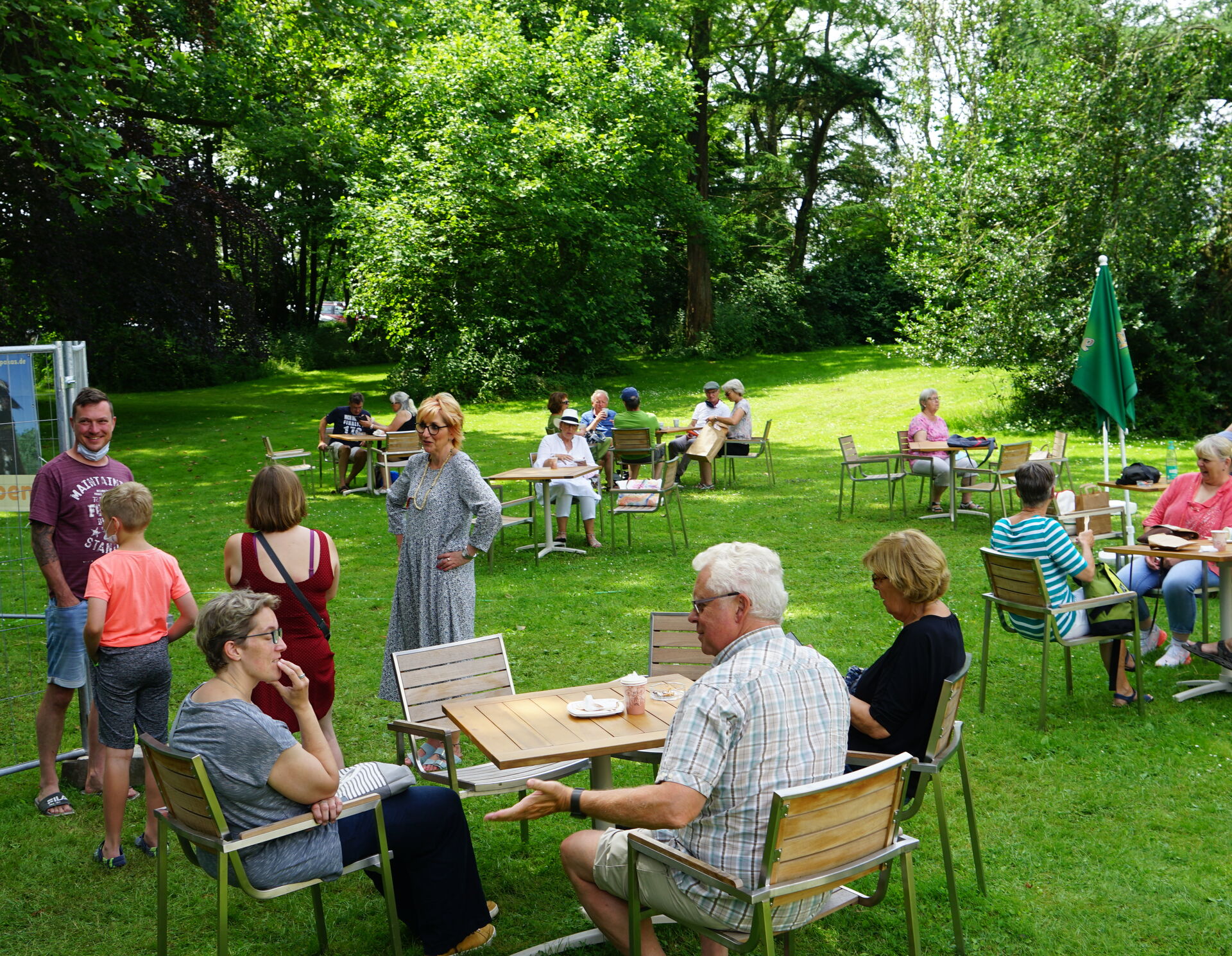 Sommermarkt, Wiese mit Tischen und Stühle, teilweise besetzt. Es sind zahlreiche Menschen auf dem Bild