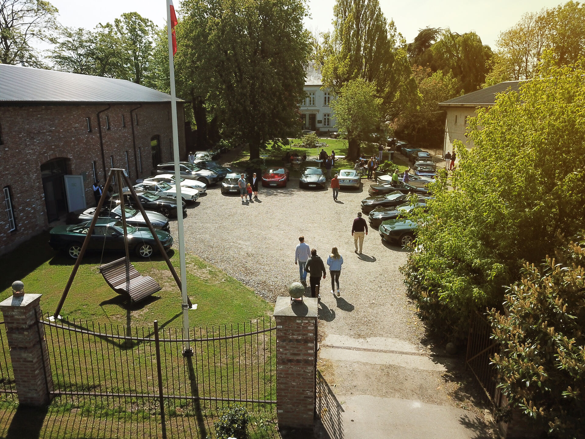 Drohnenaufnahme Einfahrt Wittkielhof, links eine Schaukel-Liege, rechts offenes Tor, Hofplatz ist voll mit Autos