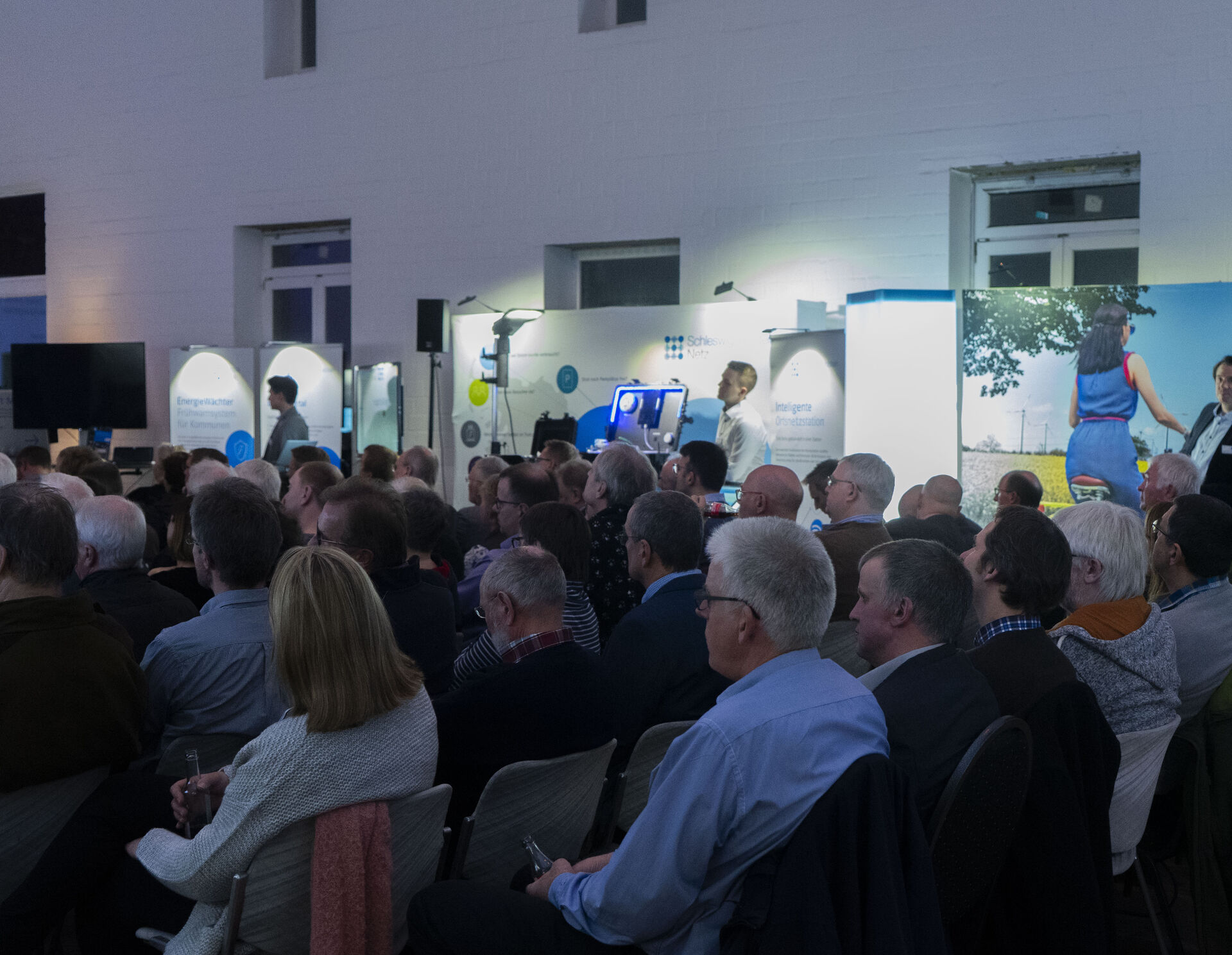 Präsentations-Stände vor einer Scheune, Sprecher und Gäste sind auf dem Bild, GFokus auf sitzende Gäste