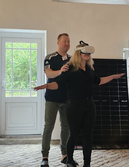 Innenraum, Frau mit VR-Brille, Mann stützt sie von hinten, Fernseher im Hintergrund