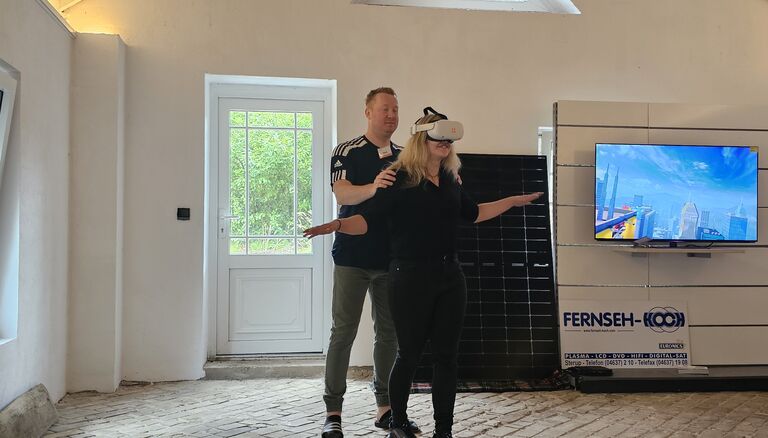 Innenraum, Frau mit VR-Brille, Mann stützt sie von hinten, Fernseher im Hintergrund