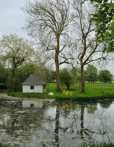 See, vorderes Ufer an unterem Bildrand, hinteres Ufer Bildmitte, dahinter weißses Häuschen am See, umgedrehtes Boot und Schild mit Schriftzug Hoffnung