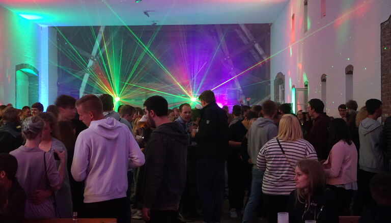 Party in einer Scheune. Raum ist voll mit Leuten. Grünes ud Violettes Licht blitzt durch die Menge.