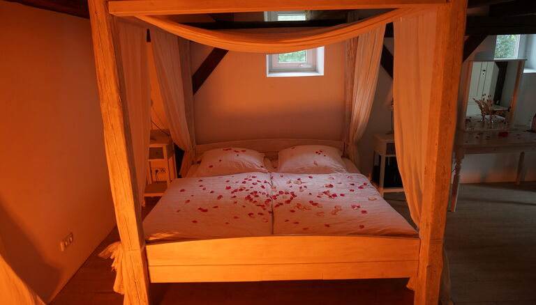 Himmelbett, betrachtet vom Fußende her. Weiße Bettwäsche, mit Rosenblättern bestreut