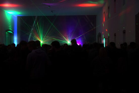Ein dunkler Raum voll mit kaum zu erkennenden Personen. Im Hintergrund befinden sich Leuchten, die grüne Lichtstrahlen aussenden.