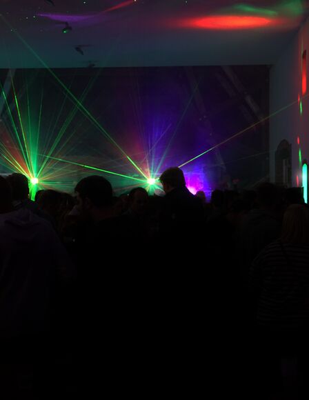 Ein dunkler Raum voll mit kaum zu erkennenden Personen. Im Hintergrund befinden sich Leuchten, die grüne Lichtstrahlen aussenden.