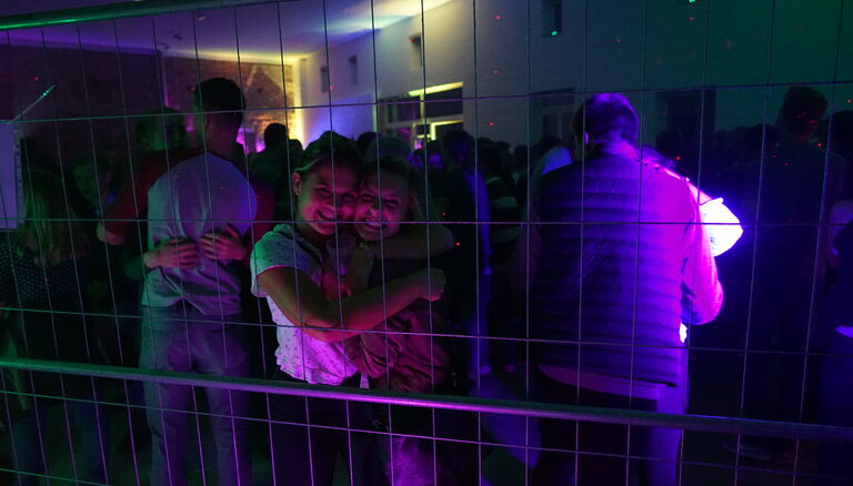 Die Scheune während eine Party im Dunkeln. Violettes, rotes und grünes Licht. Vordergrund ein Bauzaun. Dahinter zwei Mädchen, sie grinsen in die Kamera.