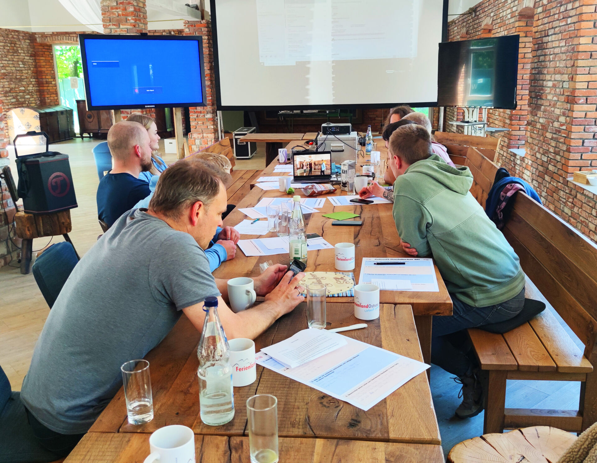 Seminar in Eventscheune, Teilnehmer sitzen an langem Tisch vor mobilen Bildschirmen. Aufgenommen von Tisch-Mitte