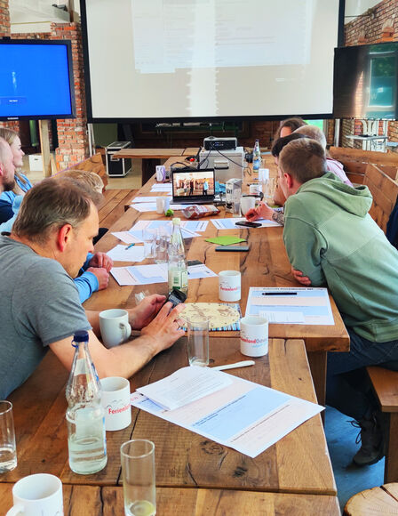 Seminar in Eventscheune, Teilnehmer sitzen an langem Tisch vor mobilen Bildschirmen. Aufgenommen von Tisch-Mitte