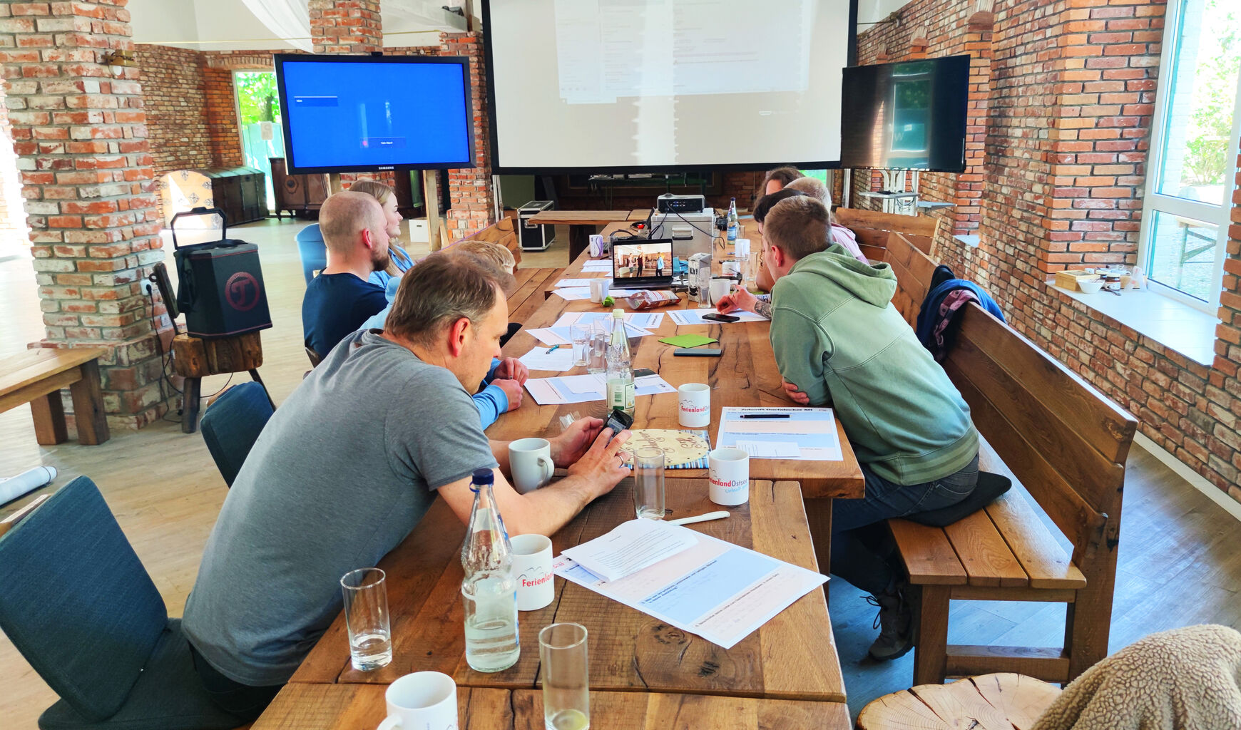 Seminar in Eventscheune, Teilnehmer sitzen an langem Tisch vor mobilen Bildschirmen. Aufgenommen von Tisch-Mitte