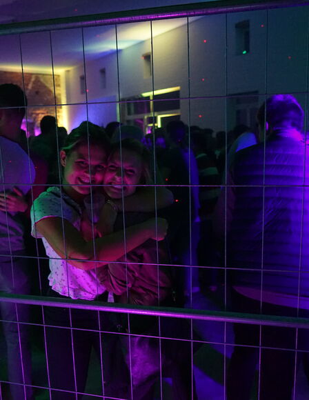 Die Scheune während eine Party im Dunkeln. Violettes, rotes und grünes Licht. Vordergrund ein Bauzaun. Dahinter zwei Mädchen, sie grinsen in die Kamera.