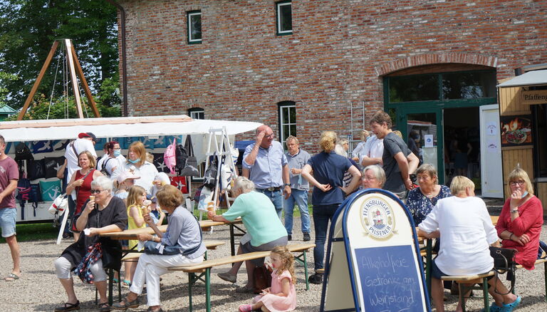 Somemrmarkt, Vordergrund, Bierzeltgarnituren, Menschen sitzen darauf und laufen drumherum, Hintergrund Scheune und Taschen-Verkaufsstand