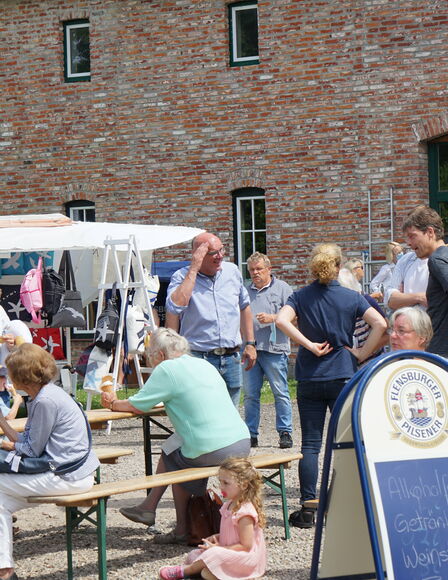 Somemrmarkt, Vordergrund, Bierzeltgarnituren, Menschen sitzen darauf und laufen drumherum, Hintergrund Scheune und Taschen-Verkaufsstand