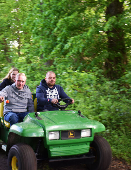 Zwei Männer und eine FRau fahren auf einem John Deere Gator über einen Waldweg