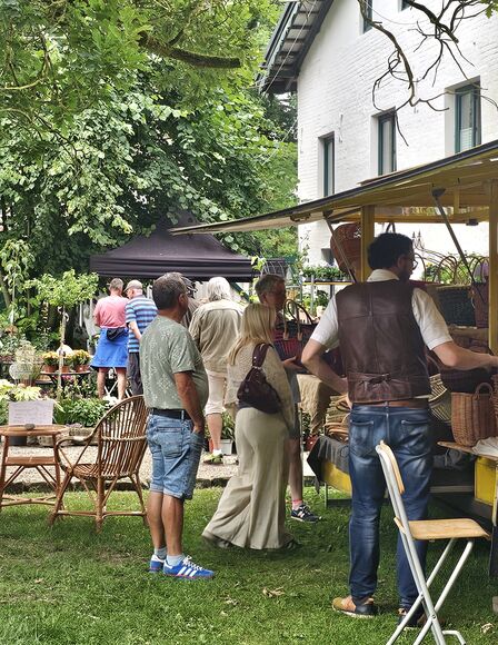 Szene Sommermarkt, rechts Stand mit Körben, Links Baum, zahlreiche Personen im Bild