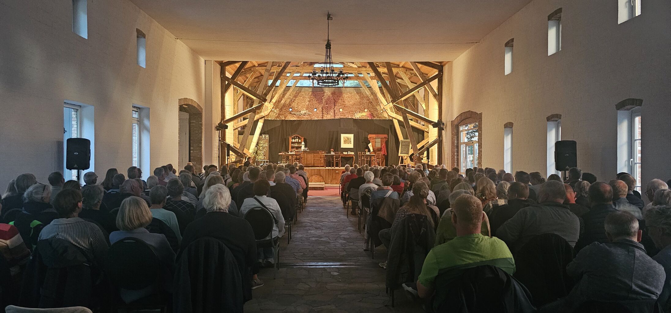 Asmus-Petersen Scheune, Hintergrund Bühne, Vordergrund und Bildmitte, Stuhlreihen gefüllt mit Menschen