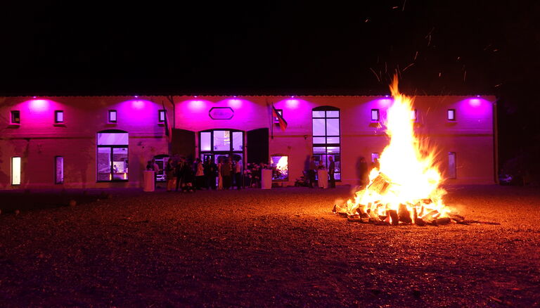 Ein Lagerfeuer brennt hoch und hell vor der Eventscheune. Die Scheune ist von Außen magentafarben beleuchtet.