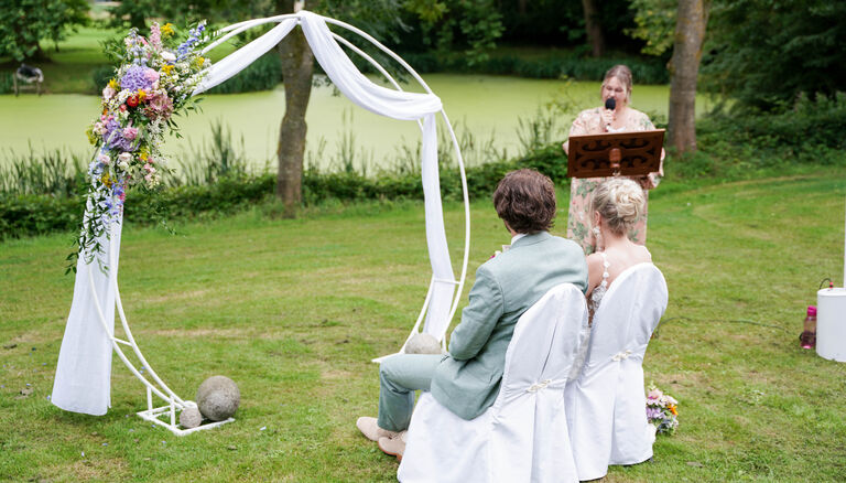 Hochzeitspaar sitzt vor Stoffbespanntem Bogen, Rücken zur Kamrea, schaut nach hinten links. Hintergrund eine Rednerin