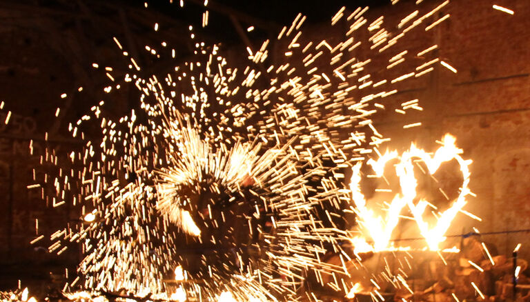 Im Vordergrund eine kreis- oder herzförmige Form aus Funken. Im Hintergrund zwei brennende, herzförmige Metallrahmen.