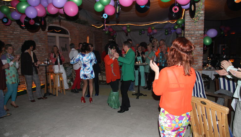 Mehrere Menschen feiern in einem Raum, unter der Decke schweben zahlreiche Luftballons.