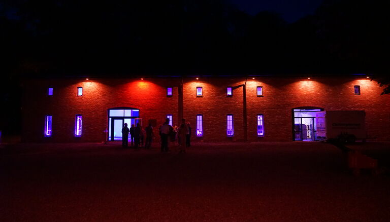 Scheune von Aussen in der Dämmerung. Aussenbeleuchtung rot-orange. Durch Fenster ist violette Innenbeleuchtung sichtbar.
