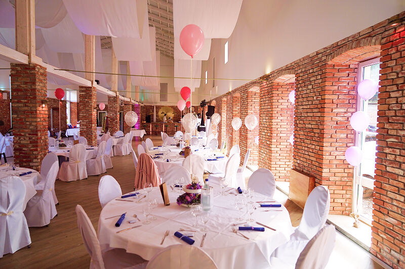 Eventscheune, Weitwinkel-Aufnahme. Weiße und rosafarbene Dekorationen. Blaue Servietten. Weiße Bezüge auf Tischen und Stühlen.