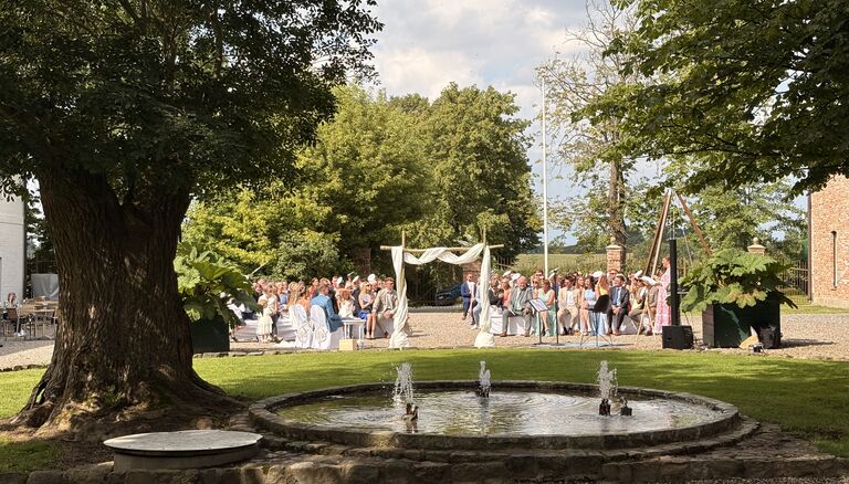 Hochzeit auf dem Hofplatz des Wittkielhofs. Im Vordergrund Bäume.