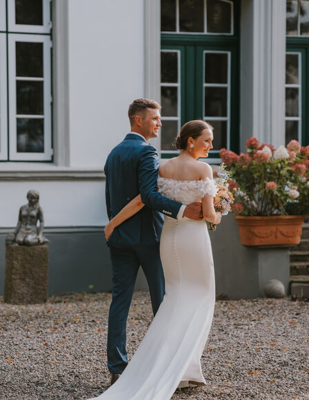 Hochzeitspaar läuft am Hauptgebäude entlang, beide schauen Richtung Kamera, laufen aber nach rechts