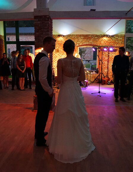 Hochzeitspaar in Eventscheune bei Dämmerung. Hochzeitsgesellschaft und -Band im Hintergrund.