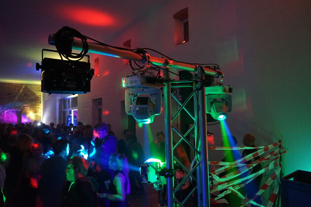 Nahaufnahme eines Gerüsts, an dem Lampen befestigt sind, die verschiedenfarbige Lichtstrahlen aussenden. Im Hintergrund sind zahlreiche Leuet zu sehen.