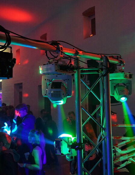 Nahaufnahme eines Gerüsts, an dem Lampen befestigt sind, die verschiedenfarbige Lichtstrahlen aussenden. Im Hintergrund sind zahlreiche Leuet zu sehen.