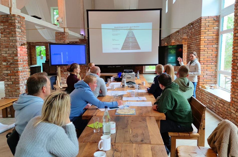 Seminar in Eventscheune, Teilnehmer sitzen an langem Tisch vor mobilen Bildschirmen. Aufgenommen vom Tischende