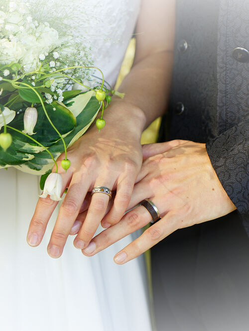 Hände von einem Hochzeitspaar. Eheringe sind zu sehen. Die Braut hält einen Strauß in der rechten Hand. Der Bildrand hat einen weißen Blur.