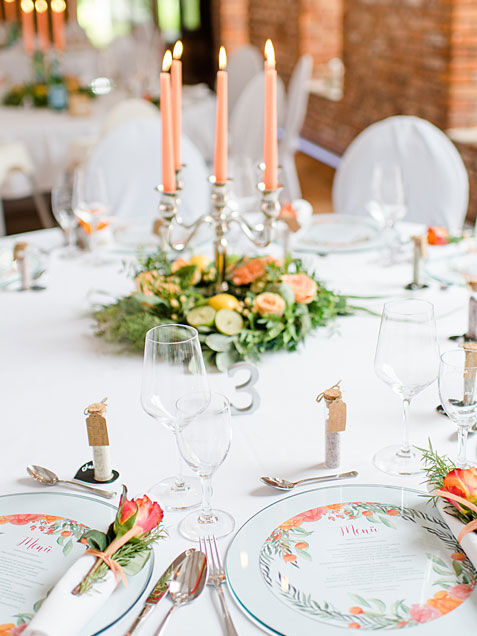 Eventscheune. Tische dekoriert mit pfirsichfabenen Kerzen. Auf den Tellern liegen Zettel, auf denen das Menü angegeben ist.