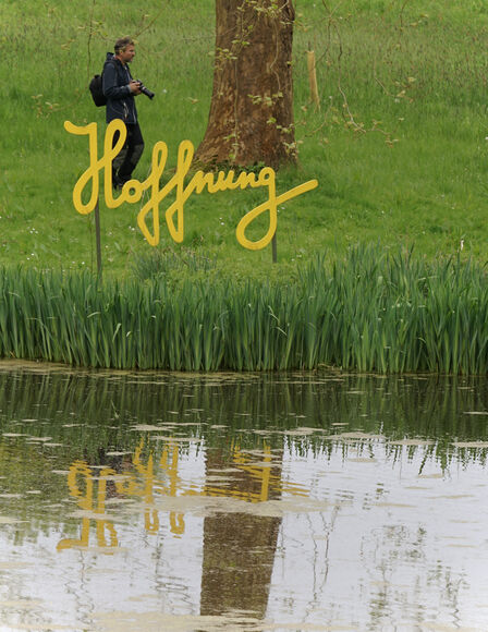 Vordergrund, See und Seeufer mit Schilf. Im Schilf steht ein Schild mit Schriftzug Hoffnung. Dahinter ist Wiese