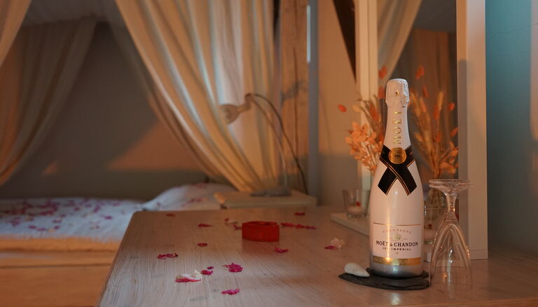 Im Hintergrund ein mit Rosenblättern dekoriertes Bett. Im Vordergrund ein Tischlein mit Sektflasche und Gläsern.