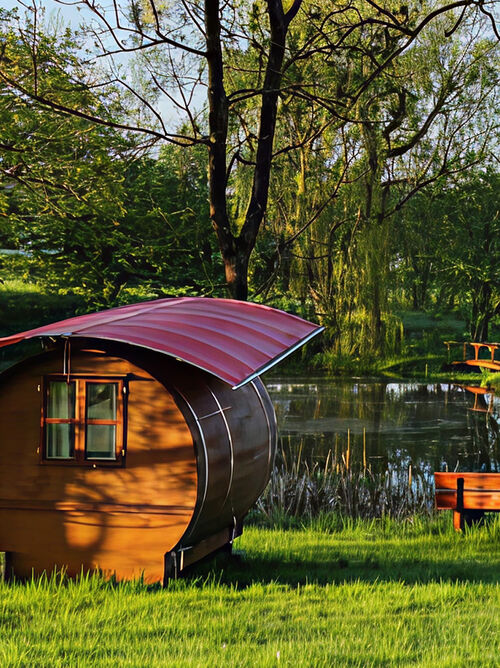 Bild vom See. Vorne links das Fass, darüber aufgespannt ist ein Dach aus roter Plane. Auf der anderen Seite des Sees steht ein weißes Häuschen.